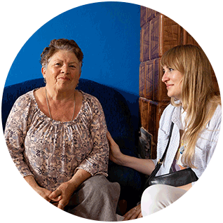 Round photo of a Ukrainian refugee and a therapist she talks to regularly through a Project-HOPE supported program. She has short hair above the ears, a slight smile and is sitting on a couch. Her therapist looks at her with a smile and pats her arm.