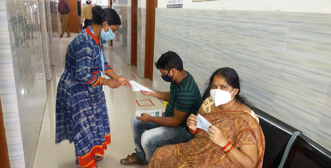 A woman in India, wearing a copper and bronze sari, smiles under a white paper mask as she holds up another protective mask. A man next to her, wearing a black mask, talks to a health care worker distributing supplies from Project HOPE.