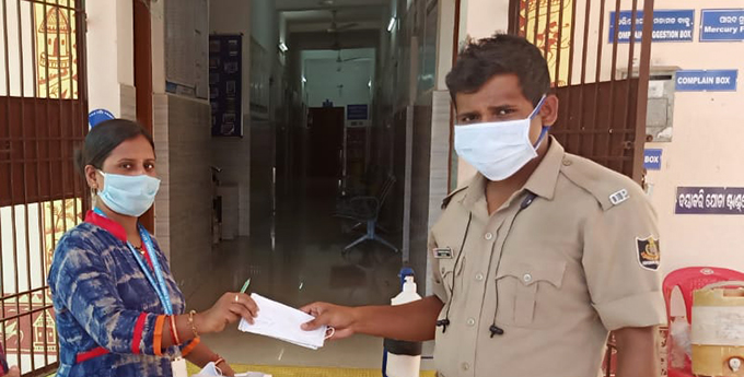 A woman in India, wearing a blue and red dress and a light blue lanyard, hands a man in a tan uniform a large stack of protective masks from Project HOPE, in front of a brown metal gate leading to a health facility.