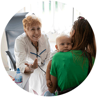 A photograph of a Project HOPE team-supported health care worker smiling and sitting in a medical office as she talks to a mom holding a small baby on her shoulder.