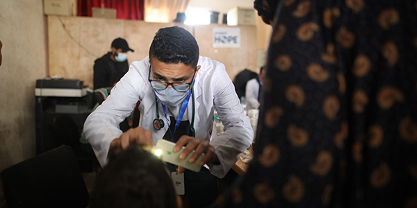 Photograph of a Project HOPE-supported community health worker in a makeshift clinic in Gaza. They are wearing a white lab coat, stethoscope and medical mask as they use a phone light to examine a child seeking health care.