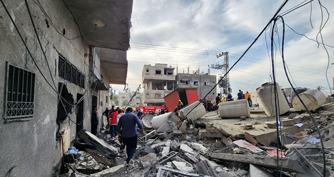 A photo of a destroyed concrete building in Rafah, Gaza shows people walking over the rubble among water tanks and hanging wires, all against a cloudy blue sky.