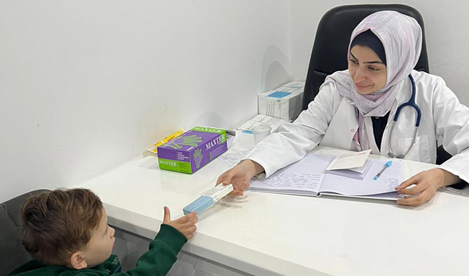 A photo from inside Gaza of a white clinic room containing a desk and two black chairs. Medicine boxes are sitting on the desktop where Dr. Maram, a Project HOPE team member, is handing a small boy a box as she smiles down at him.