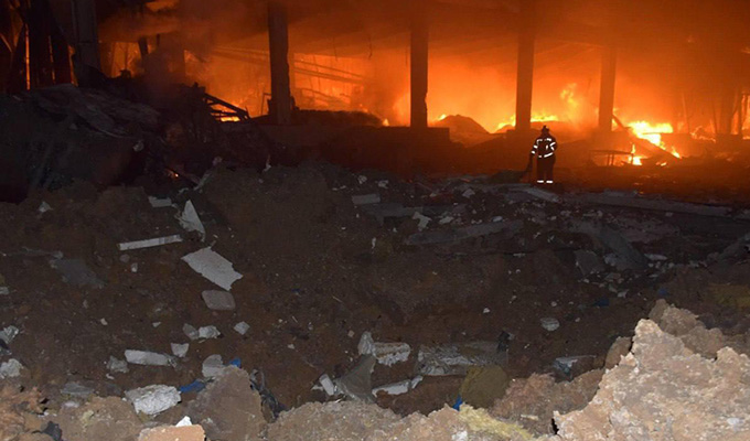 A photo of a destroyed warehouse and rubble against the background of a fire and a firefigher whose outline is barely visible except for the relfective stripes on his suit.