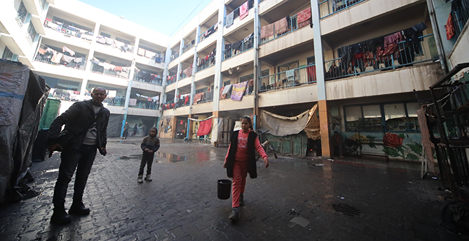 Photograph from a center for displaced families in Rafah a family in winter clothes including a man and two children. One of the children carries a bucket of war as they all look at the camera.