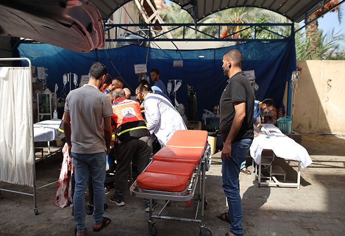 A photograph of an outdoor treatment room in Gaza with many health workers crowded around a patient. IVs, gurneys, tarps and sheets surround them.