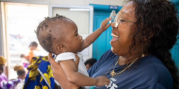 Photograph of a Project HOPE-supported community health worker in Sierra Leone holding a small baby in a tshirt and earrings. The baby is grabbing the woman's glasses as the woman laughs, against the background of a community clinic.