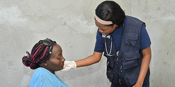 A photograph of a Project HOPE supported health worker wearing medical gloves, tshirt, vest and stethoscope as she examines the side of a woman's face at a mobile medical clinic in Haiti.