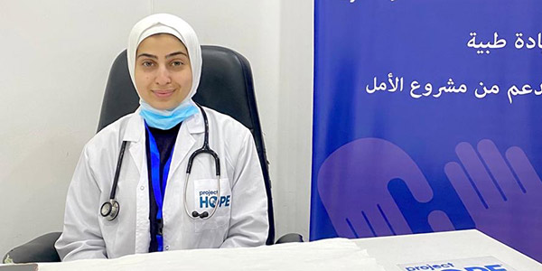 Photograph of a Project HOPE supported health worker who is sitting at a desk in a medical office at our primary health care clinic in Gaza. She is smiling at the camera as she wears a lab coat, stethoscope and hair covering.