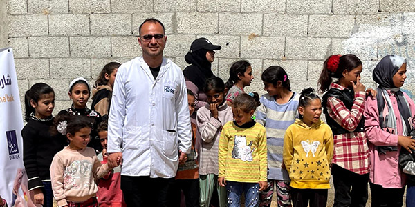 Photograph of a Project HOPE supported health worker is standing with a group of various ages of children. He is smiling at the camera as he wears a doctor's coat with Project HOPE logo, glasses and dark pants as a leader of children's activities in Gaza.