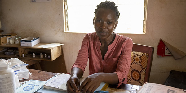 A photograph of a Project HOPE supported midwife in Sierra Leone where she is sitting at the desk in her office and looking softly, but intently at the camera. The chair she sits in is patterned and her desk is covered in medical documents and medicines with a window directly behind them.