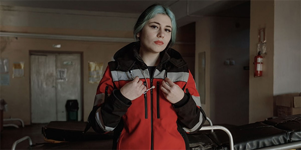 Photograph from Ukraine shows a Project HOPE supported ambulance driver in her gear and stethoscope looking at the camera with intensity. She is in a darkened room of a medical clinic with a fire extingusiher on the wall in the background.