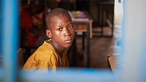 A photograph of a boy in a classroom looking pensively through the bars of a school window. He wears a yellow t-shirt and sits slightly slouched forward.