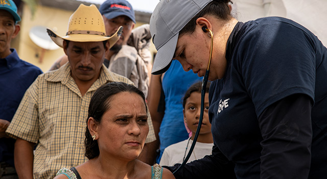 A photograph of Project HOPE health worker in Ecuador as they use a stethoscope to check the health of a woman who is a migrant.