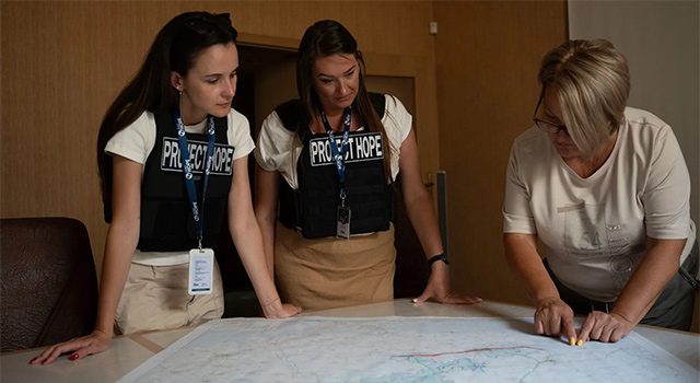 A photograph of a Project HOPE mobile team in Ukraine where three health workers read a map in a wood-paneled room as they plan a response.