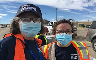 Photo of two health care workers in orange traffic vests and navy caps as they stand in a parking lot.