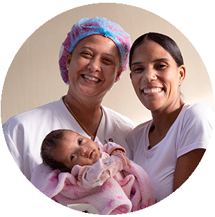 A photograph showing two health workers in white scrubs smiling brightly as they hold a small baby in a pink blanket at a Project HOPE-supported hospital in the Dominican Republic.