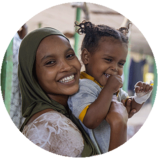 A photograph of a young mother in Ethiopia smiling as she holds her laughing toddler at a Project HOPE-supported clinic. She wears a heasd scarf and tshirt while her daughter wears a yellow polo under a grey shirt.