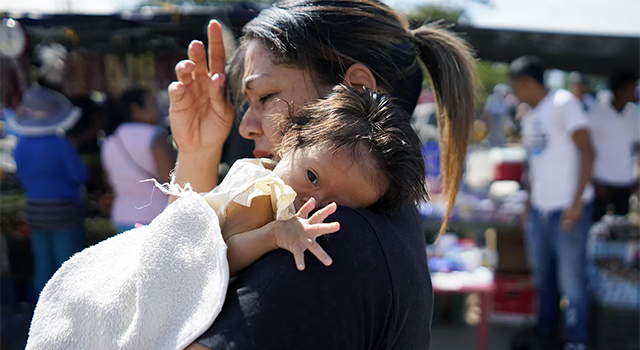 A woman in Colombia looks to the left shielding her eyes and her young baby's eyes from the bright sun light. The baby reaches her tiny hand towards the viewer from under a white blanket.