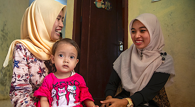 Two Indonesian women wearing light-colored clothing and head scarves smile brightly at each other in room with a wooden door. A young baby sits on one woman's lap. She wears a red shirt with 3 cats on it as she looks quizzically at the camera.