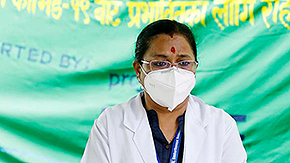 A health worker in Nepal wears a white medical face mask, glasses, and a white lab coat, as well as a navy shirt and lanyard. She looks down sadly in front of a green, yellow and blue banner about COVID relief, reading in English: Supported by Project HOPE.