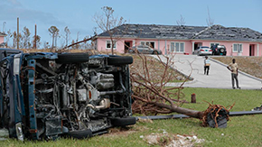 A landscape with a blue sky shows a wrecked car on its side alongside many upended signs and trees. A pink house and its concrete driveway are in the background with 2 distant figures looking in the distance.