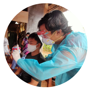 A health worker in Indonesia wears a face shield, white medical mask, gown and gloves as he takes the temperature of a young man wearing a navy shirt and white mask.