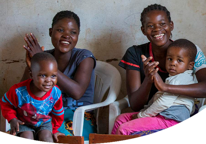 Two sets of mother and child sit in white plastic chairs in Malawi. They are smiling and clapping.