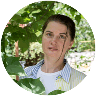 A round cutout of a Ukrainian women who is wearing a color-blocked oxford shirt and standing next to tall plants and trees at a community center in Poland.