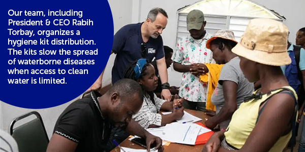 Photograph of Project HOPE staff and people from the local community interacting over a desk where hygiene kits are being distributed inside a white room with a large window.