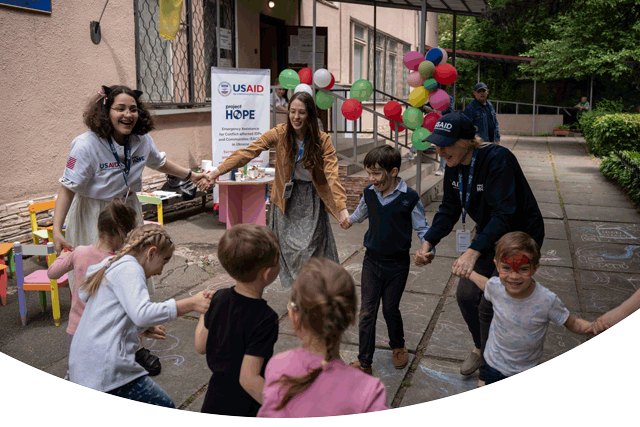 A photograph shows group of smiling and laughing teachers and children holding hands as they move in a circle. They play a game in an outside courtyard decorated with ballons. They are part of a Project HOPE-supported mental health program for children traumatized by the war in Ukraine.